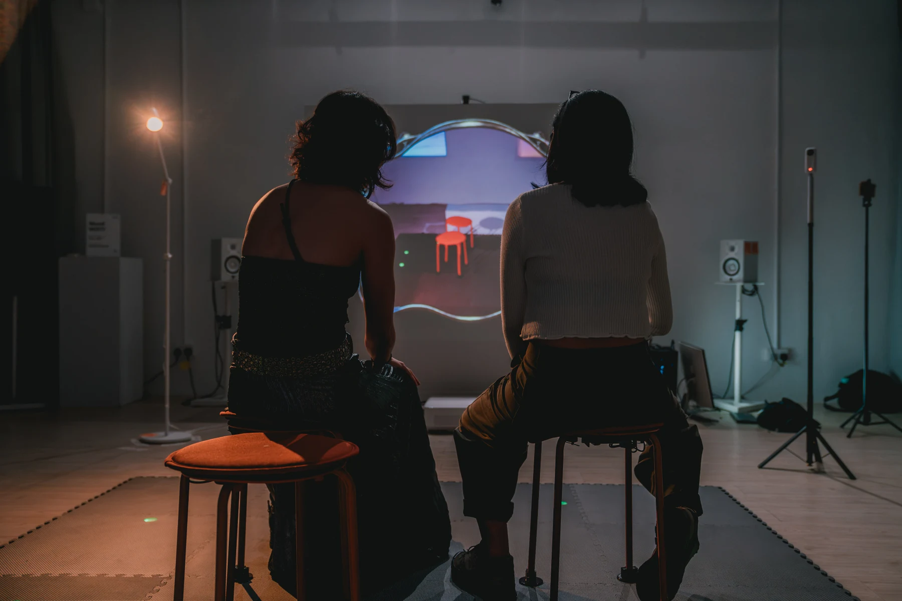 Two people sit facing a projected view of the room that shows the stools without them.